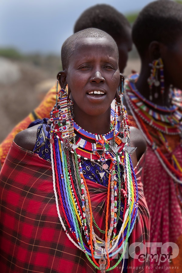 African Safari - Kenya. . . . . Closeup of young Masai woman in bright red shuka, and beaded jewellery, with other women,  Masai village, Kenya, Africa