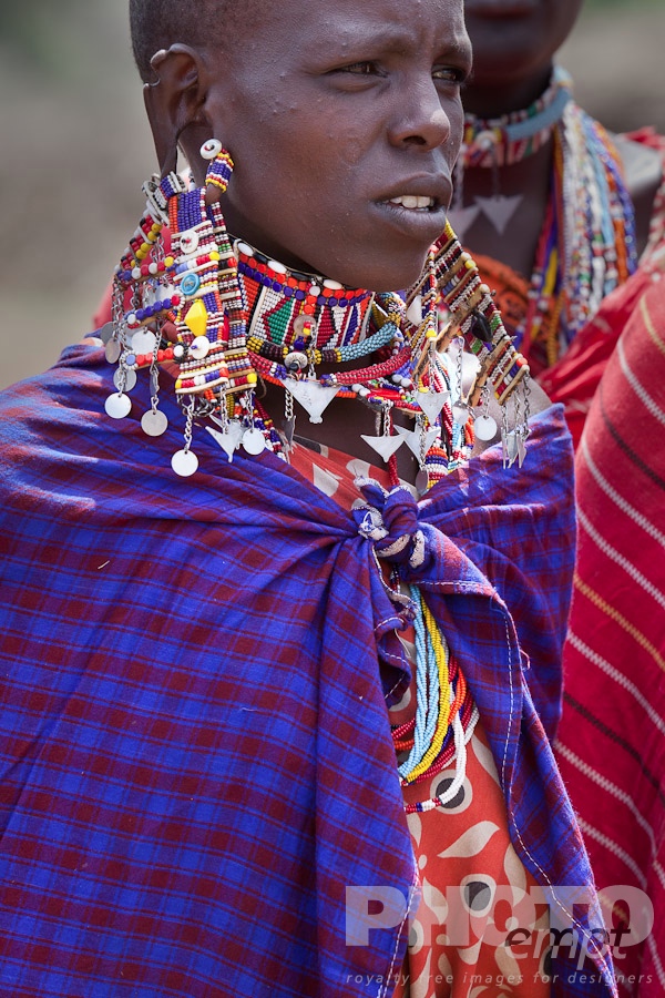 African Safari - Kenya. . . . . Closeup of young Masai woman in blue and red shuka, and beaded jewellery, with other women,  Masai village, Kenya, Africa