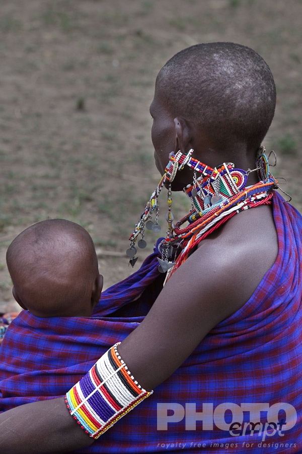 African Safari - Kenya. . . . . Young Masai woman dressed in blue and red traditional shuka, holding a baby, back view, sitting, Masai village, Kenya, Africa