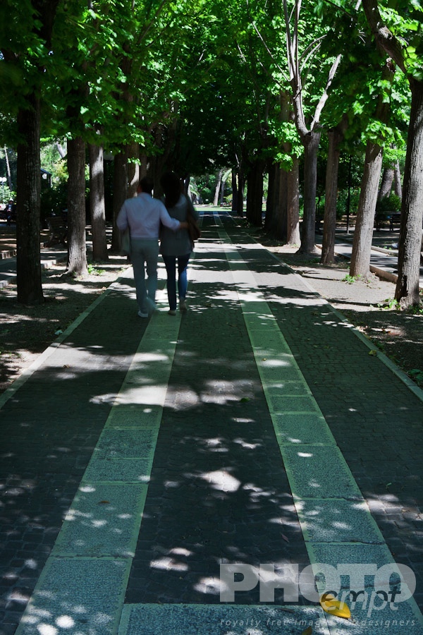 Italy South - Puglia. . . . . Back view of male and female couple with motion blur, walking in park, avenue lined by trees, paved with white lines, dappled sunlight, Lecce,  Puglia, Southern Italy