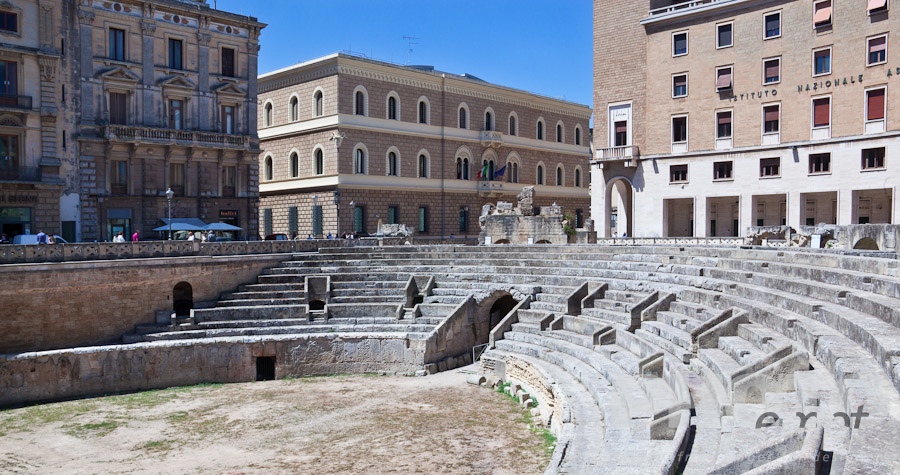 Italy South - Puglia. . . . . Roman amphitheatre with semicircular arena in foreground; shops, Renaissance, Baroque and modern facades beyond, in morning sunshine with blue sky, Lecce, Salento, Puglia, Southern Italy