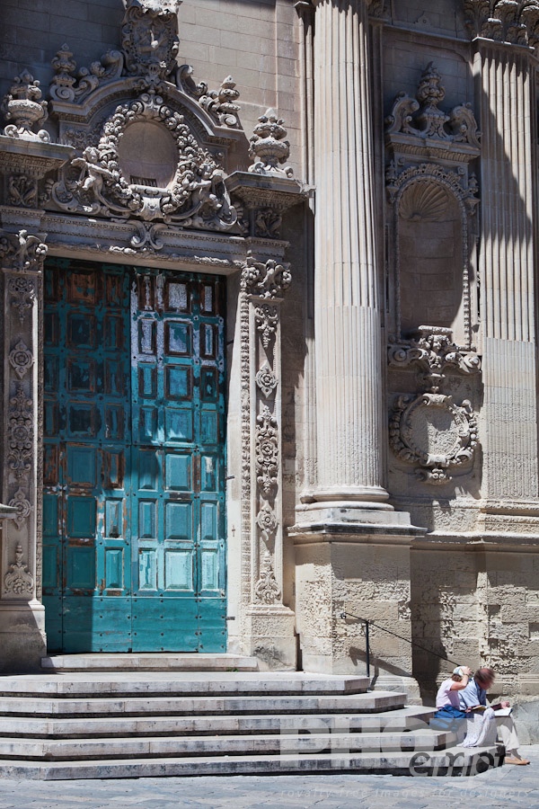Italy South - Puglia. . . . . Close up of doorway, Baroque ornamentation and blue faded paintwork of door, church of Santa Chiara, with tourist couple sitting on steps, looking at guide book, sunny spring morning, Lecce, Salento, Puglia, Southern Italy