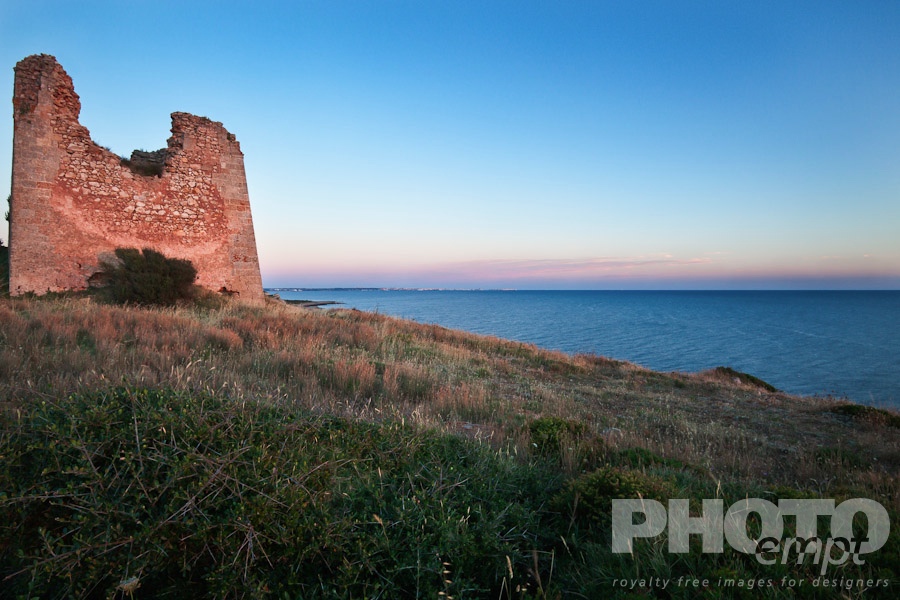 Italy South - Puglia. . . . . Medieval defense tower overlooking coastline, calm sea, at sunset with pink light on stone tower, and pink clouds, Gallipoli town on horizon,  Salento, Puglia, Southern Italy