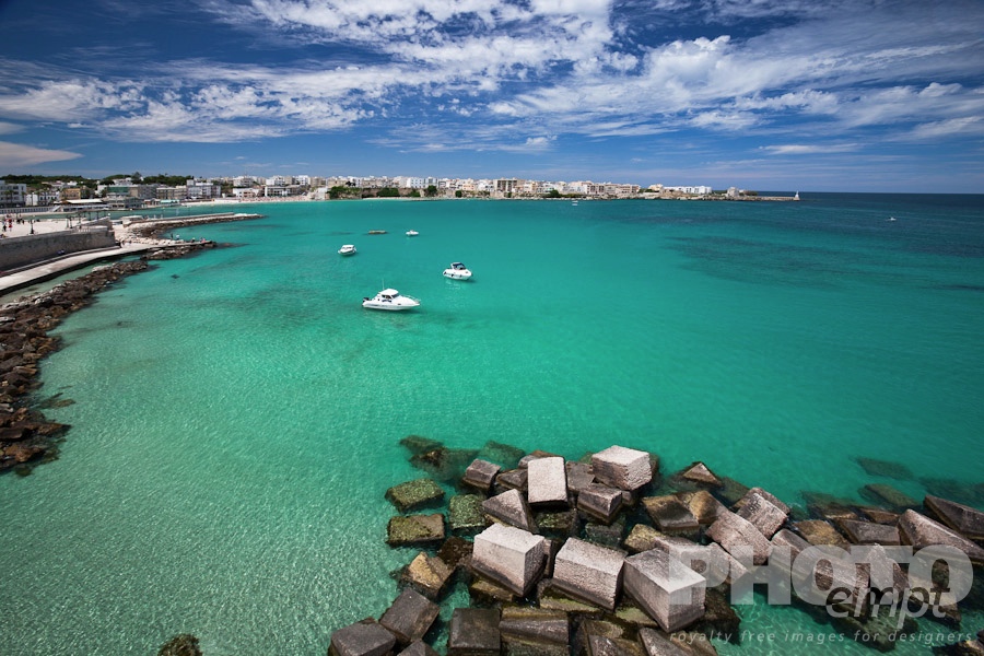 Italy South - Puglia. . . . . View of Otranto harbour with clear green water, moored sailboats and motorboats, medieval fortifications and town beyond, sunny spring morning with dramatic clouds in blue sky, Salento, Puglia, Southern Italy