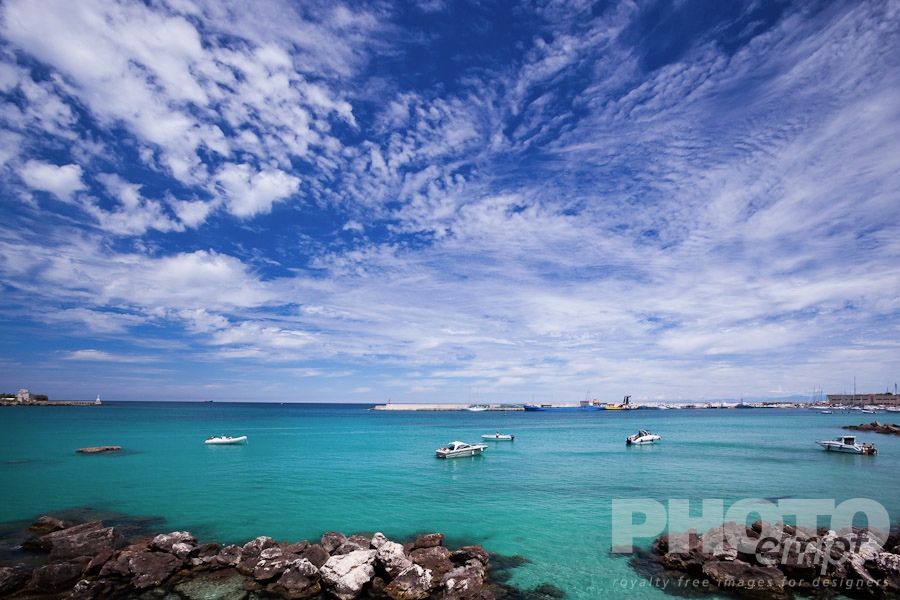 Italy South - Puglia. . . . . View of Otranto harbour with clear green water, moored sailboats, cargo boat, and motorboats, sunny spring morning with dramatic clouds in blue sky, Salento, Puglia, Southern Italy