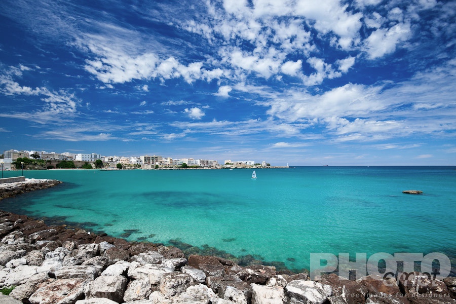 Italy South - Puglia. . . . . View of Otranto harbour with clear green water, moored sailboats and motorboats,  town beyond, sunny spring morning with dramatic clouds in blue sky, Salento, Puglia, Southern Italy