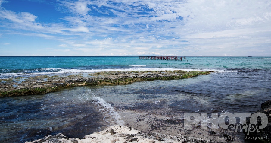 Italy South - Puglia. . . . . Rocky beach with bright blue water, red metal pier, cargo boat on horizon, and snow capped mountains of Albania visible across sea, dramatic clouds in blue sky, Salento, Puglia, Southern Italy