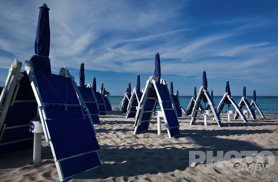 Italy South - Puglia. . . . . Close up of rows of folded blue beach chairs and umbrellas casting long shadows across fine sandy beach, blue calm sea and dramatic clouds in sunny blue sky, Salento, Puglia, Southern Italy