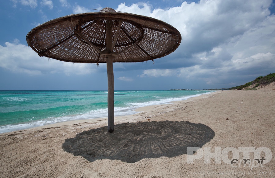 Italy South - Puglia. . . . . Empty, fine sandy beach , with single wicker beach umbrella casting shadow; white foam, green sea colour, blue sunny sky, with storm threatened,  Salento, Puglia, Southern Italy