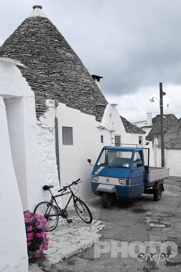 Italy South - Puglia. . . . . Street view: town of Alberobello, World Heritage site, with trullo houses, white painted stone with piled stone conical roofs, paved street and red flowers, bicycle and blue traditional van set against monochrome view,  Puglia, Southern Italy