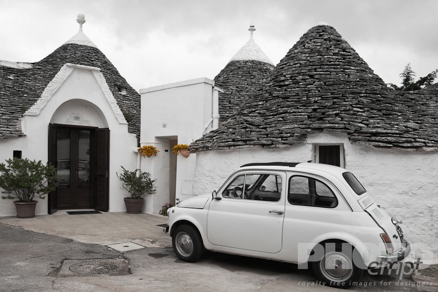 Italy South - Puglia. . . . . Street view: town of Alberobello, World Heritage site, with trullo houses, white painted stone with piled stone conical roofs, paved street, yellow flowers, white traditional car set against monochrome view,  Puglia, Southern Italy