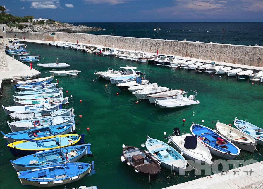 Italy South - Puglia. . . . . Aerial view of colourful fishing boats moored in sheltered harbour, vivid green clear water, blue sea and sky beyond, sunny spring afternoon, Salento, Puglia, Southern Italy
