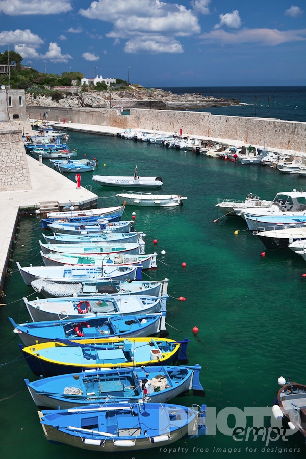 Italy South - Puglia. . . . . Aerial view of colourful fishing boats moored in sheltered harbour, vivid green clear water, blue sea and sky beyond, sunny spring afternoon, Salento, Puglia, Southern Italy
