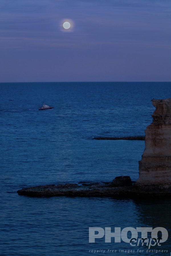 Italy South - Puglia. . . . . Cliffs and rocky coastline of Salento at twilight, with full moon reflection on sea, carabinieri motor boat in distance, Puglia, Southern Italy