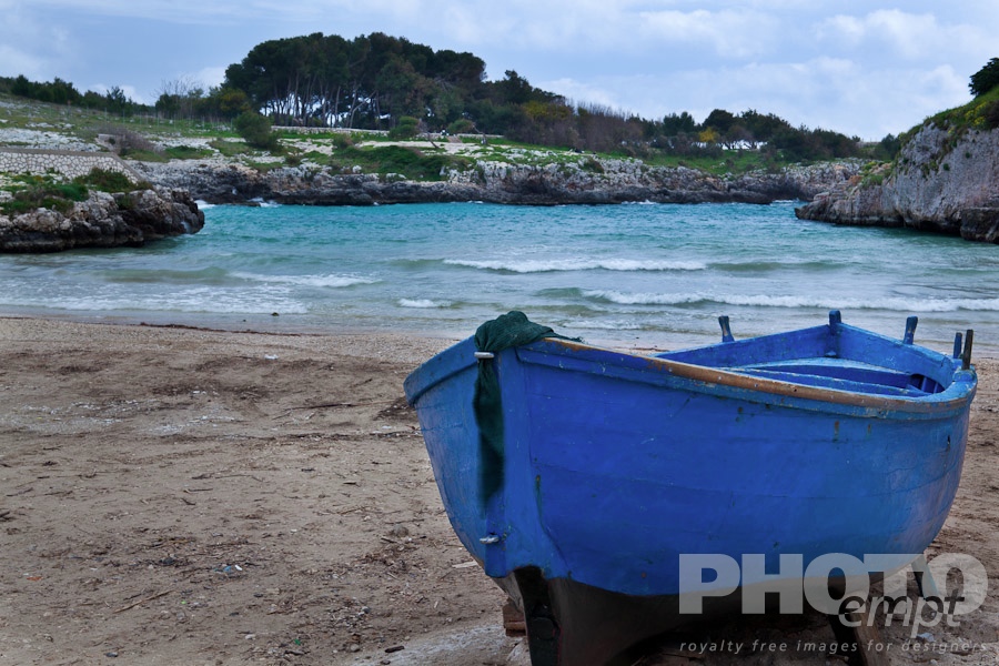 Italy South - Puglia. . . . . Beached blue wooden fishing boat on sandy shore of Porto Badisco inlet, with rocky shore line, blue sky, Salento, Puglia, Southern Italy