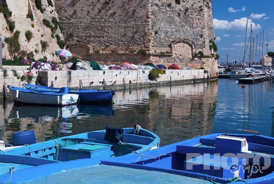 Italy South - Puglia. . . . . Wooden fishing boats moored in harbour, with fishing nets on quayside below the medieval walls of Gallipoli, and sailboats beyond, sunny spring morning, Salento, Puglia, Southern Italy