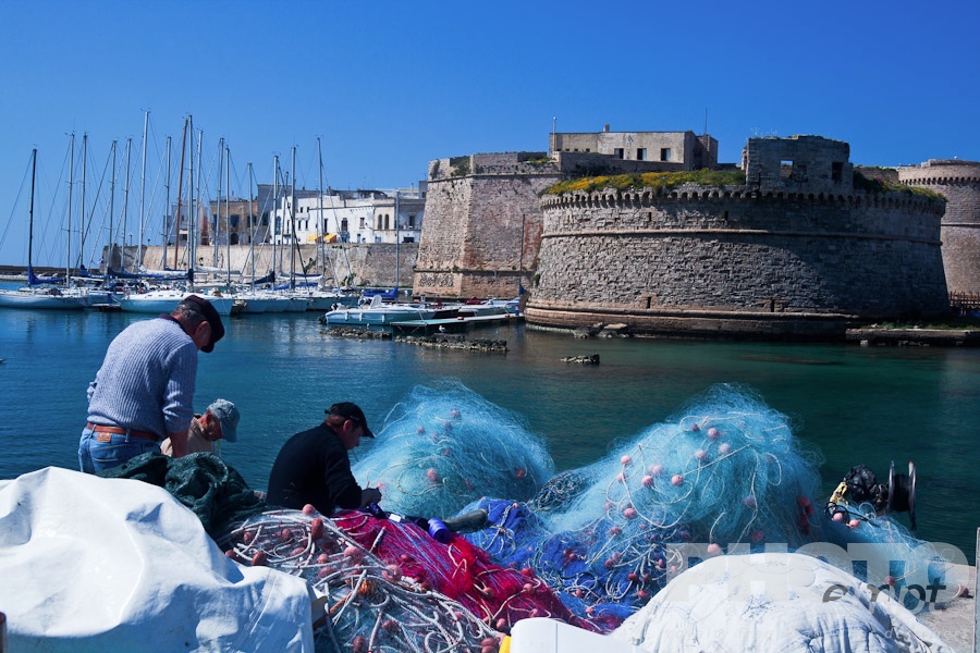Italy South - Puglia. . . . . Fishermen mending colourful nets in harbour with sailboats moored beneath medieval stone walls of Gallipoli, sunny spring morning with blue sky, Salento, Puglia, Southern Italy