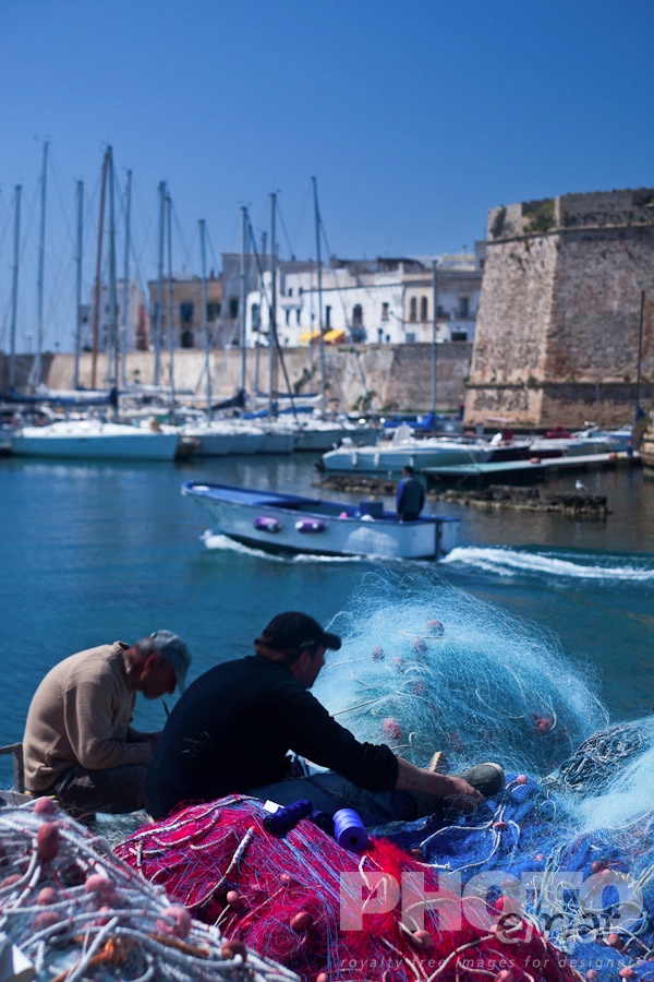 Italy South - Puglia. . . . . Fishermen mending colourful nets in harbour with sailboats moored beneath medieval stone walls of Gallipoli, sunny spring morning with blue sky, Salento , Puglia, Southern Italy