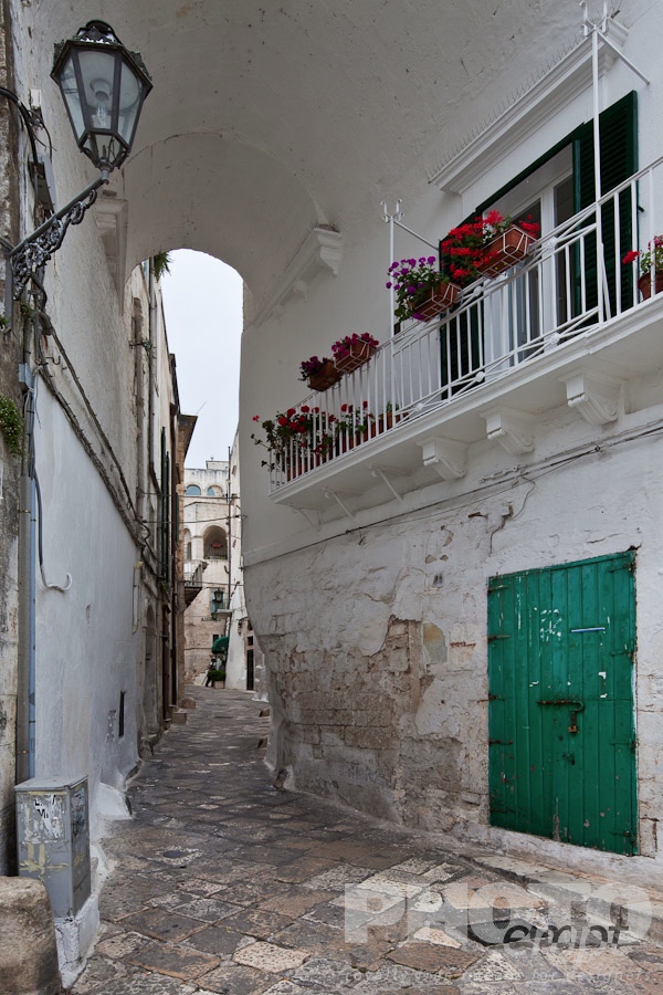 Italy South - Puglia. . . . . Street in Ostuni, with typical white stucco walls, stone paving, wrought iron lamp, and archway, with flowers decorating, spring afternoon, Salento, Puglia, Southern Italy