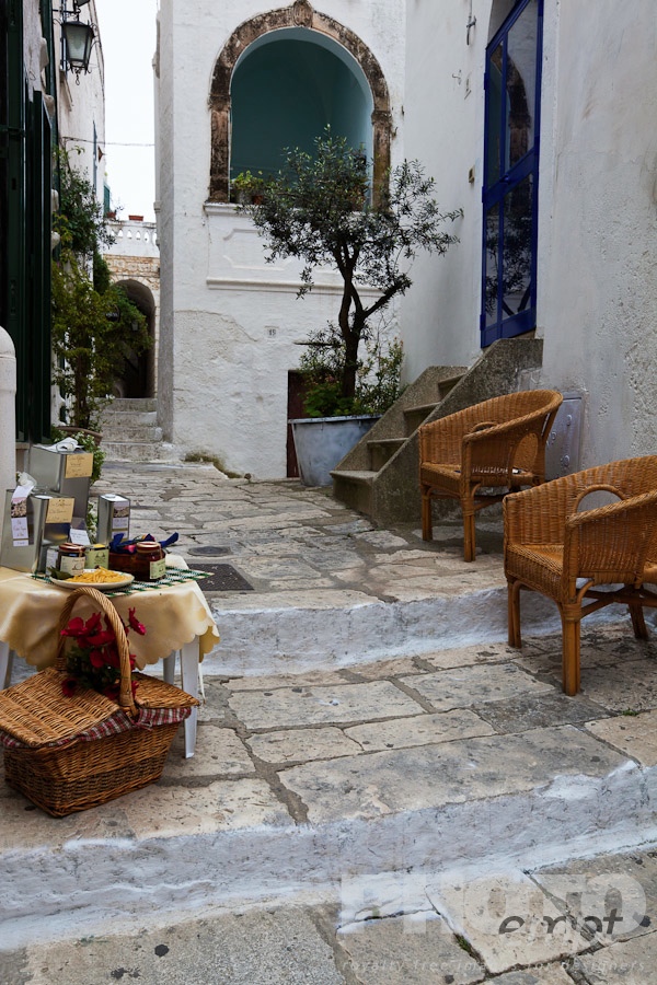 Italy South - Puglia. . . . . Street in Ostuni, with typical white stucco walls, stone paving, wrought iron lamp, and archway, with flowers decorating, and display of produce on wicker tables and chairs, spring afternoon, Salento, Puglia, Southern Italy