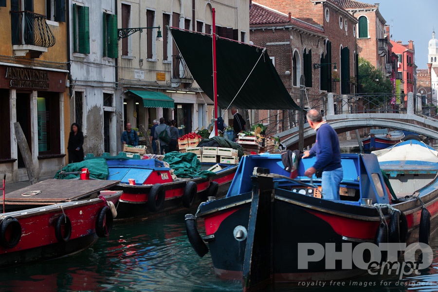 Venice in Autumn. . . . . Fruit and vegetable market on colourful blue and red barge with motion blur of vendor and shoppers, with second barge moving in foreground, sunny autumn morning in Venice, Northern Italy