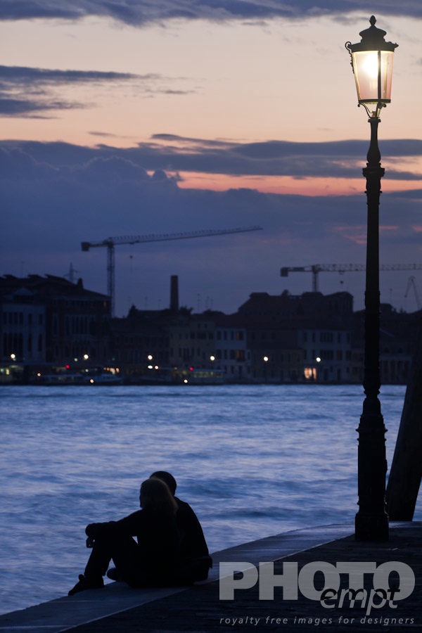Venice in Autumn. . . . . View of Canale della Giudecca, with young couple silhouetted, sitting beside illuminated wrought iron lamp on quayside, looking towards Giudecca, twilight, autumn in Venice, Northern Italy