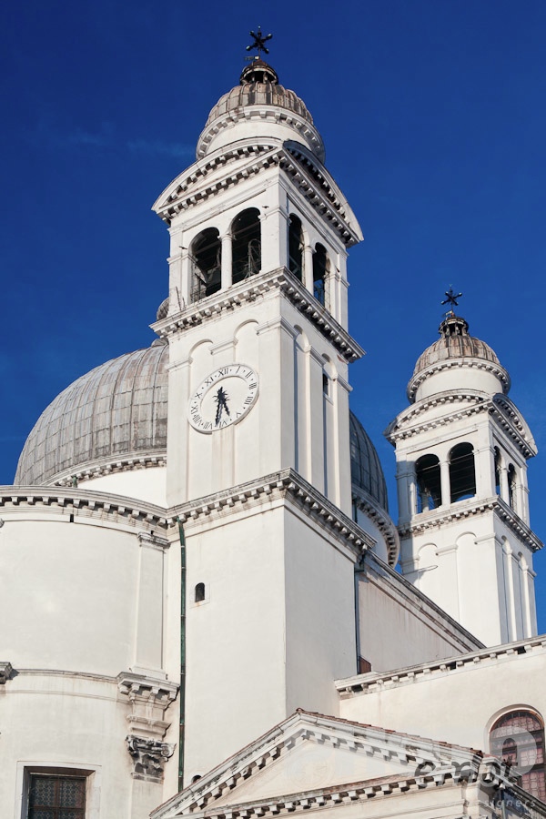 Venice in Autumn. . . . . Detail of church of Santa Maria della Salute: dome with ornamental stone scroll work and statues below, bell and clock towers, against deep blue sky, late afternoon in autumn, Venice, Northern Italy